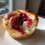 A close-up of a baked brie bite showing melted brie cheese and bright red jam inside flaky pastry.