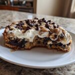 A close-up of a thick slice of chocolate chip cookie cake recipe topped with white frosting and extra chips.