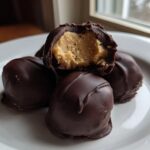 Four chocolate-covered peanut butter balls stacked on a white plate, one is broken open revealing the creamy filling.