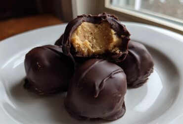 Four chocolate-covered peanut butter balls stacked on a white plate, one is broken open revealing the creamy filling.