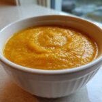 A close-up of a white bowl filled with thick, vibrant orange pumpkin soup, ready to eat.