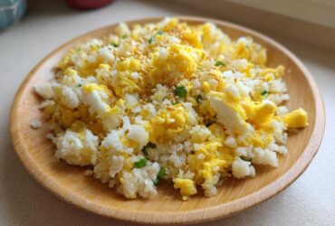 A close-up of amazing cauliflower fried rice mixed with scrambled egg, topped with sesame seeds, served on a wooden plate.