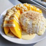 A plate of mango sticky rice featuring bright yellow mango slices, sticky white rice, and drizzled with sweet coconut cream, topped with sesame seeds.