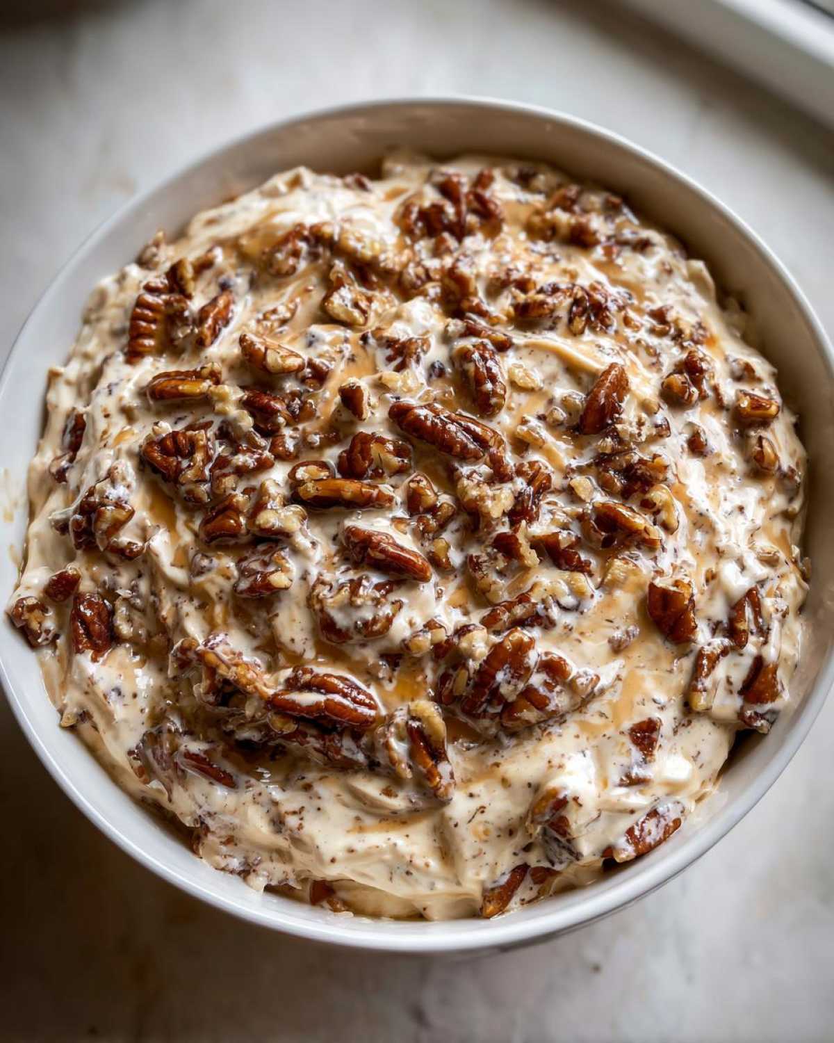Close-up of creamy pecan pie dip in a white bowl, generously topped with toasted pecans and drizzled caramel sauce.