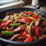 Close-up of savory pepper steak stir-fry with beef strips, red and green bell peppers, and onions in a dark skillet.