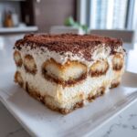 A perfect slice of homemade tiramisu recipe showing layers of cream and coffee-soaked ladyfingers, topped with cocoa powder.