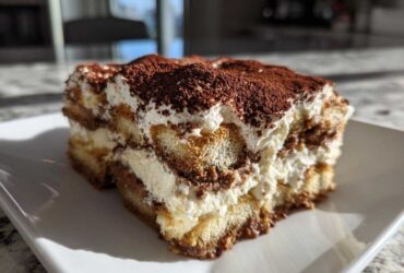 A close-up shot of a rich slice of tiramisu recipe, layered with cream and dusted heavily with cocoa powder.