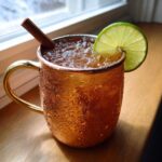 A condensation-covered copper mug filled with an apple cider moscow mule, garnished with a lime wheel and cinnamon stick.