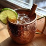 A chilled apple cider moscow mule garnished with a lime wheel and cinnamon stick in a copper mug.
