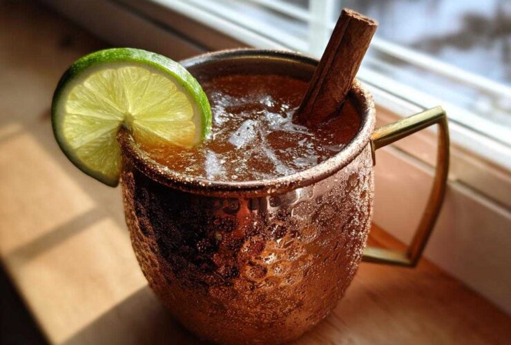 A chilled apple cider moscow mule garnished with a lime wheel and cinnamon stick in a copper mug.