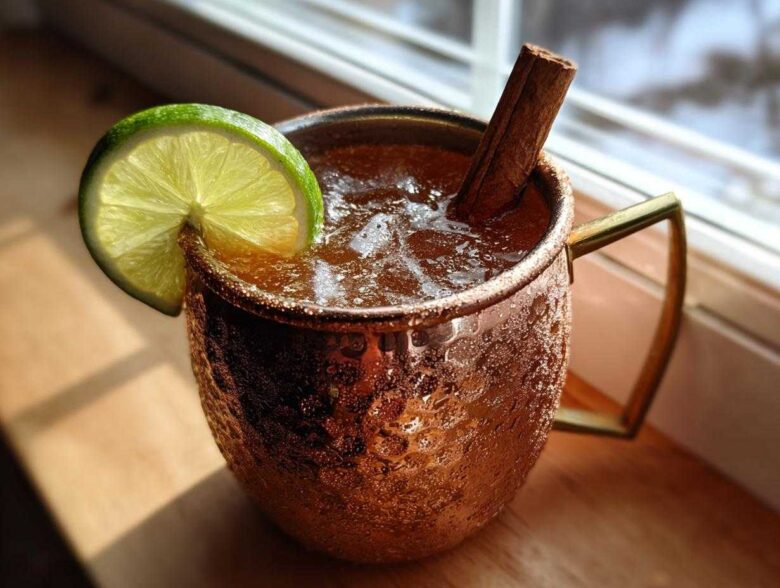 A chilled apple cider moscow mule garnished with a lime wheel and cinnamon stick in a copper mug.