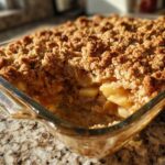 Close-up of a golden brown apple crisp recipe baked in a clear glass dish, showing the crunchy topping and soft apples.