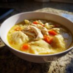 Close-up of a white bowl filled with rich, yellow broth, tender shredded chicken, carrots, and soft dumplings in chicken and dumpling.