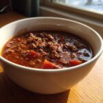 Close-up of a white bowl filled with rich, savory best chili recipe, sitting on a wooden table near a window.