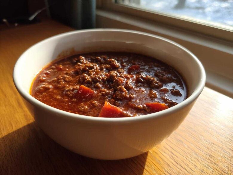 Close-up of a white bowl filled with rich, savory best chili recipe, sitting on a wooden table near a window.