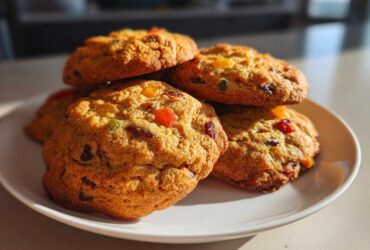 A close-up stack of freshly baked fruitcake cookies studded with colorful candied fruit pieces, sitting on a white plate.