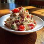 A mound of creamy blt chicken salad topped with crispy bacon pieces and bright red cherry tomatoes in a white bowl.