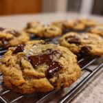 Close-up of a gooey brown butter chocolate chip cookie topped with flaky sea salt on a cooling rack.