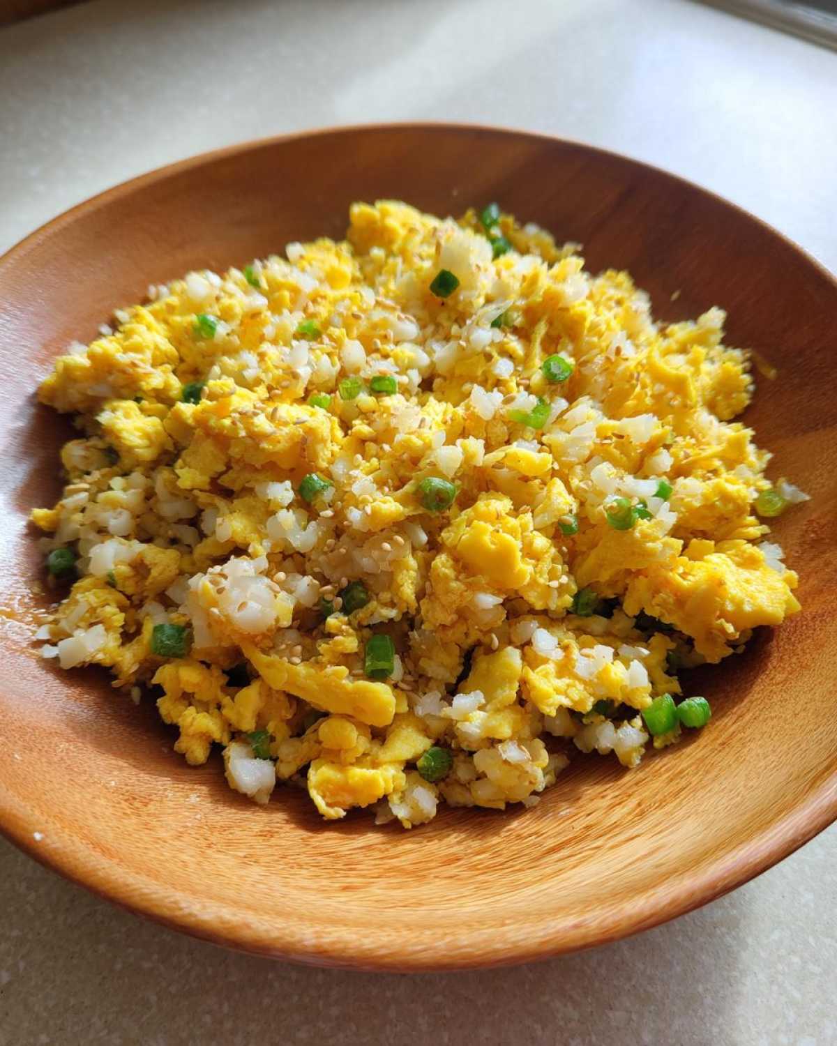 A wooden bowl filled with bright yellow cauliflower fried rice mixed with scrambled egg pieces and green onions.