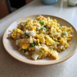 A close-up of a serving of cauliflower fried rice mixed with scrambled egg, green peas, and sesame seeds on a light wooden plate.