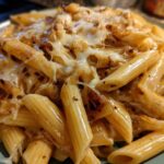 A close-up shot of penne pasta coated in a rich sauce and topped with melted, gooey cheese, characteristic of french onion pasta.