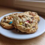 Three chewy kitchen sink cookies loaded with candy, chocolate, and pretzels on a small white plate.