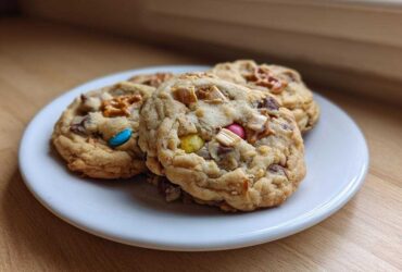 Three chewy kitchen sink cookies loaded with candy, chocolate, and pretzels on a small white plate.