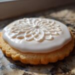 Close-up of a round, scalloped sugar cookie topped with smooth white cookie icing featuring a piped snowflake design.