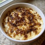 Close-up of a creamy, baked caramelized onion dip topped generously with dark, sweet caramelized onions in a white bowl.