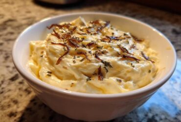 Close-up of creamy caramelized onion dip topped with crispy onions and herbs in a white bowl.