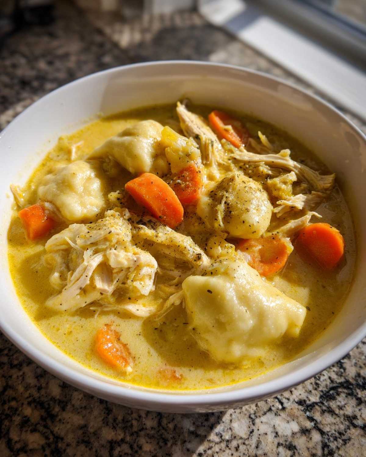 Close-up of a white bowl filled with creamy chicken and dumpling soup, featuring shredded chicken, carrots, and dumplings.