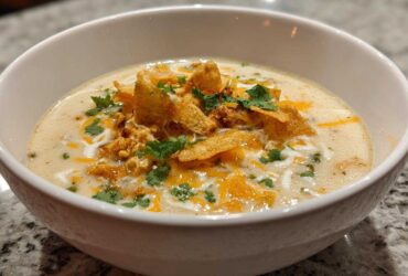 A close-up of a white bowl filled with creamy chicken tortilla soup, garnished with tortilla strips, cheese, and cilantro.
