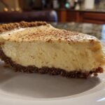 A close-up of a thick slice of creamy eggnog pie with a dark graham cracker crust, dusted with nutmeg.