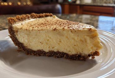 A close-up of a thick slice of creamy eggnog pie with a dark graham cracker crust, dusted with nutmeg.
