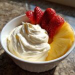A white bowl filled with creamy fruit dip, garnished with sliced strawberries and pineapple wedges.