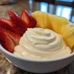 A white bowl filled with creamy fruit dip, surrounded by sliced fresh strawberries and pineapple wedges.