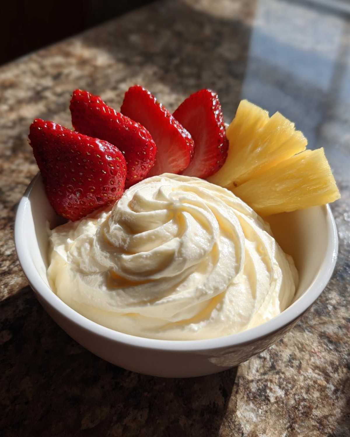 A bowl of creamy fruit dip topped with sliced strawberries and pineapple wedges.