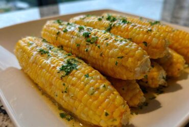 Several bright yellow ears of cooked corn covered in melted honey butter and fresh parsley.