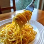 A fork lifting a twirled portion of bright yellow Creamy Lemon Pasta from a white plate.