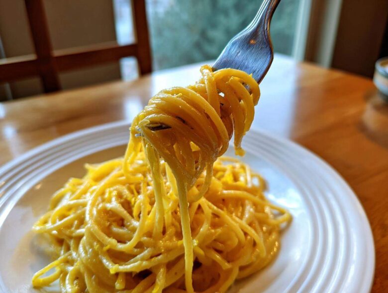 A fork lifting a twirled portion of bright yellow Creamy Lemon Pasta from a white plate.