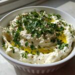 A white bowl filled with creamy white olive dip, drizzled with olive oil and topped generously with fresh chopped parsley.