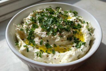 A white bowl filled with creamy white olive dip, drizzled with olive oil and topped generously with fresh chopped parsley.