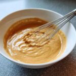 A close-up of creamy, light brown peanut sauce being whisked in a white bowl.