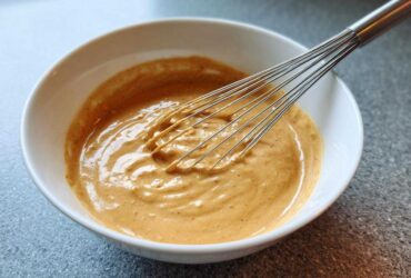 A close-up of creamy, light brown peanut sauce being whisked in a white bowl.