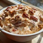 A close-up of rich, creamy pecan pie dip topped generously with whole pecans, served in a white bowl.