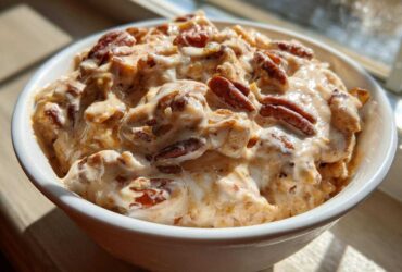 A close-up of rich, creamy pecan pie dip topped generously with whole pecans, served in a white bowl.