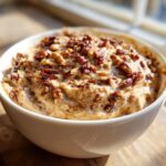 A close-up of creamy pecan pie dip served in a white bowl, generously topped with chopped, toasted pecans.