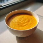 A close-up of a white bowl filled with vibrant orange, creamy pumpkin soup, lightly seasoned with black pepper.