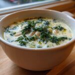 Close-up of a white bowl filled with creamy Tuscan white bean soup, topped with dark green kale and olive oil.