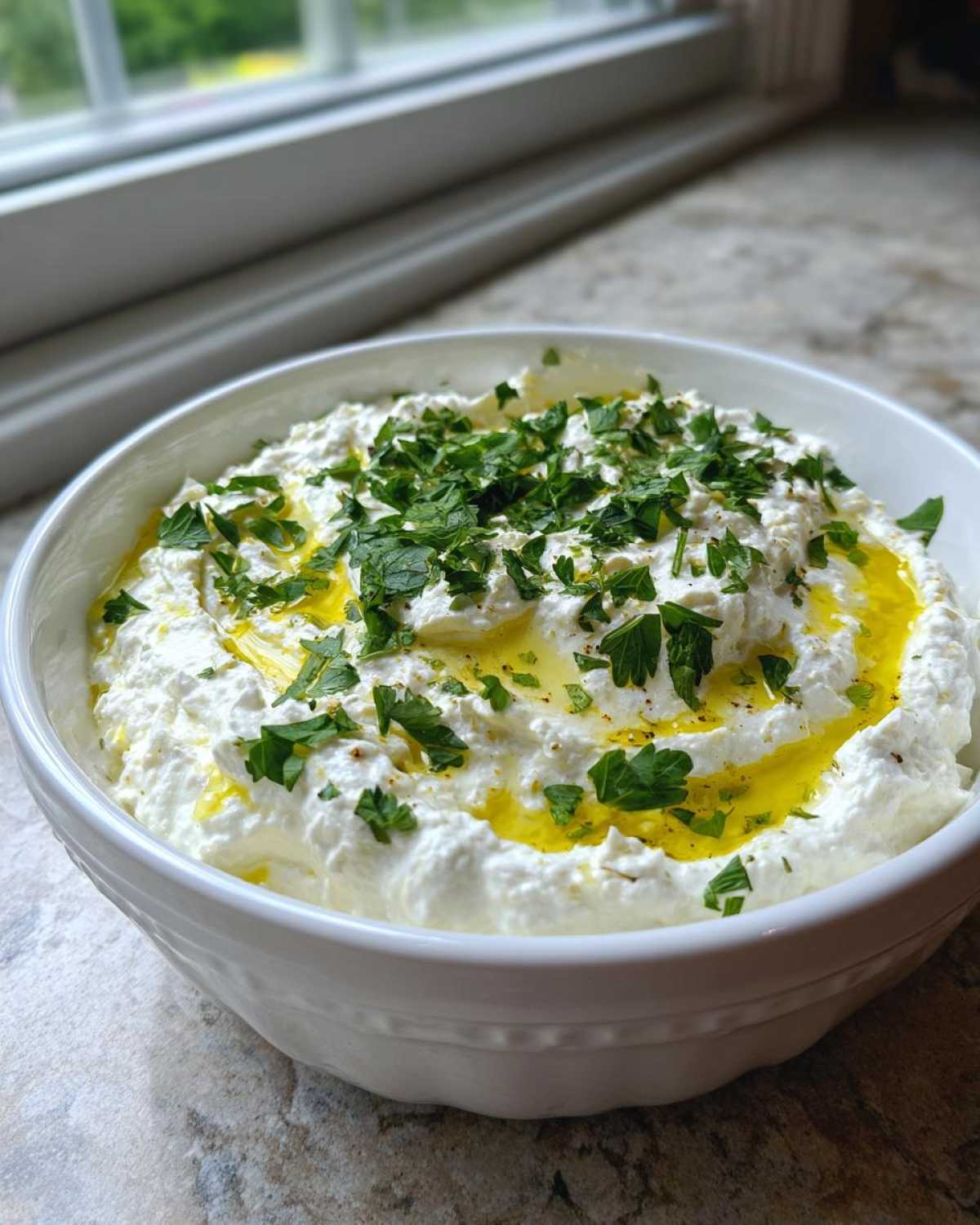 A white bowl filled with creamy white olive dip, drizzled with olive oil and topped with fresh chopped parsley.